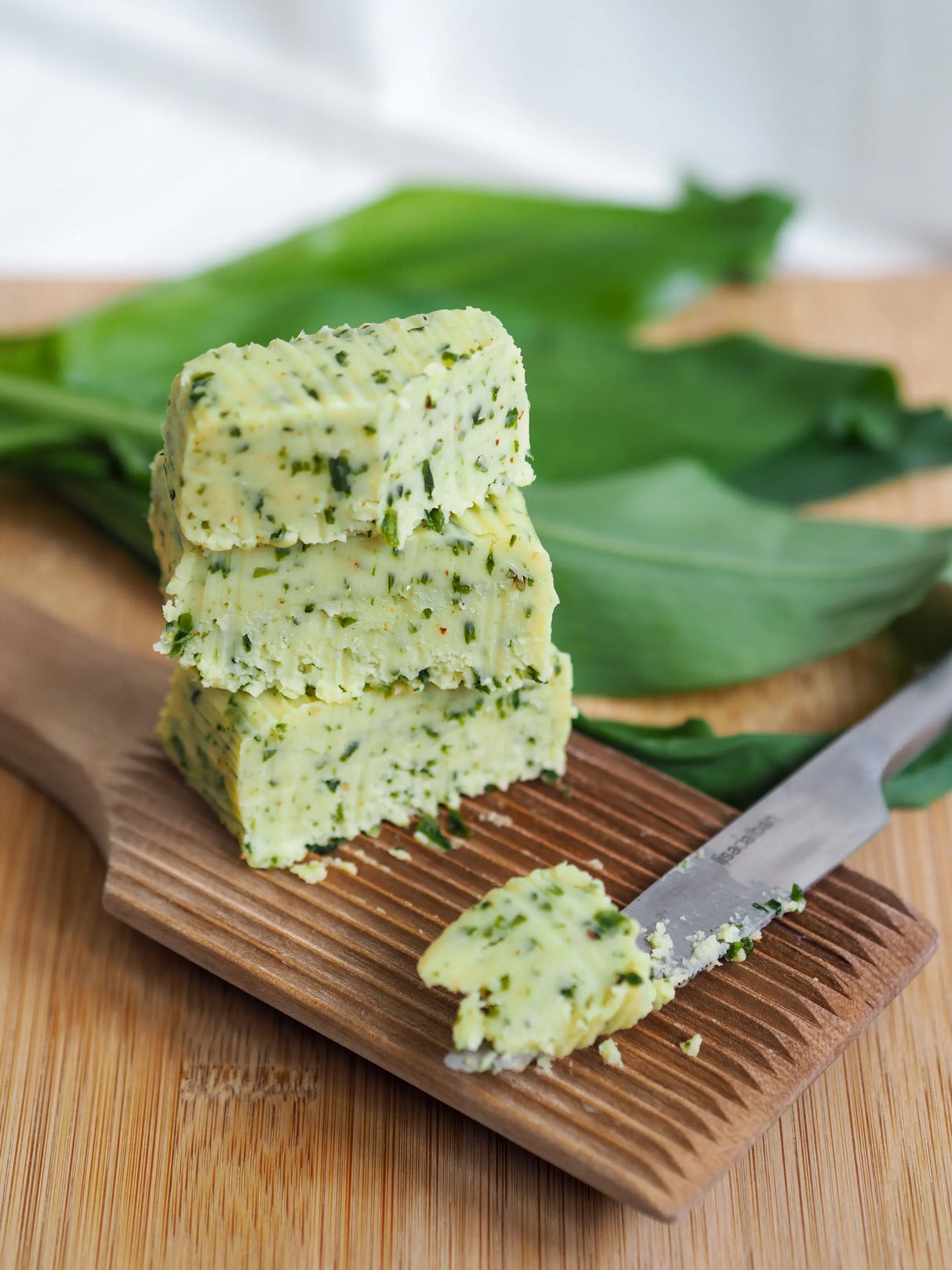 Herb-infused butter slices on wooden board with knife, fresh greens background