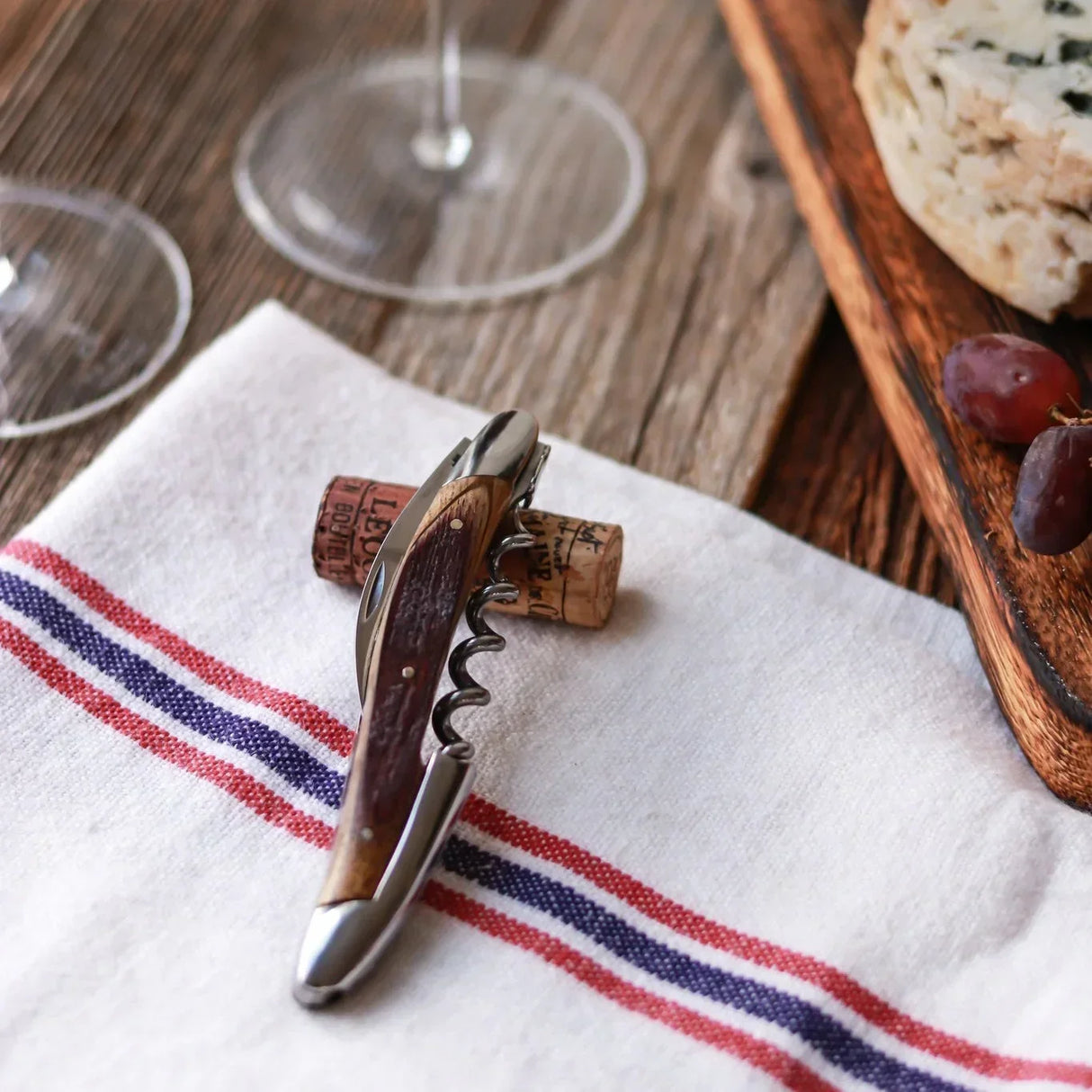 Wood-handled corkscrew on striped napkin with wine corks, grapes, cheese, and wine glasses — artisan French dining essentials