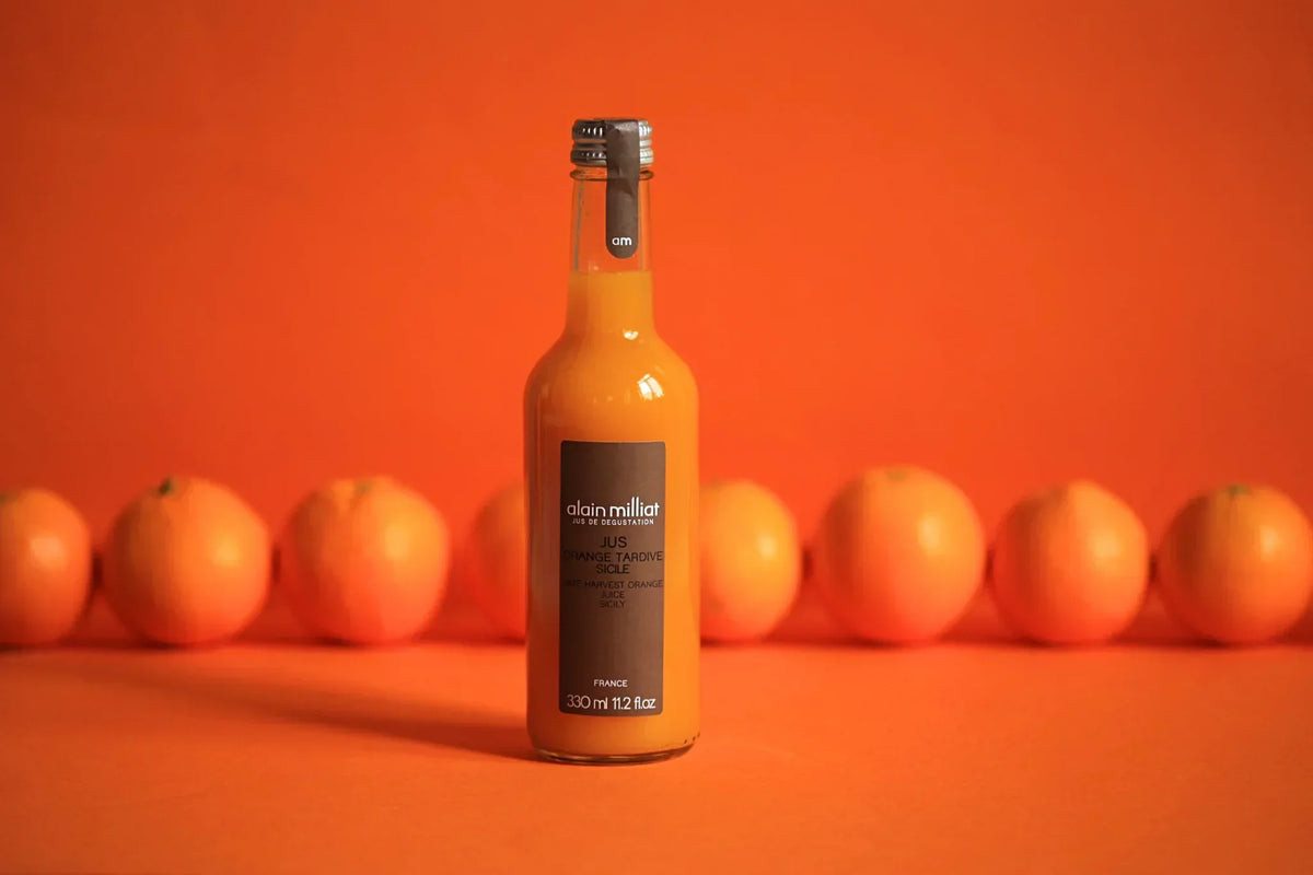 Alain Milliat Sicilian orange juice bottle with fresh oranges against vibrant orange background