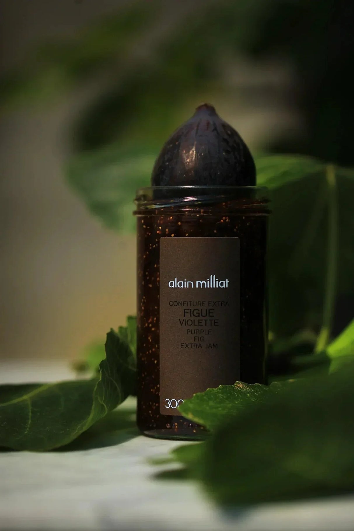 Alain Milliat purple fig extra jam in glass jar with fresh fig on top, surrounded by green leaves