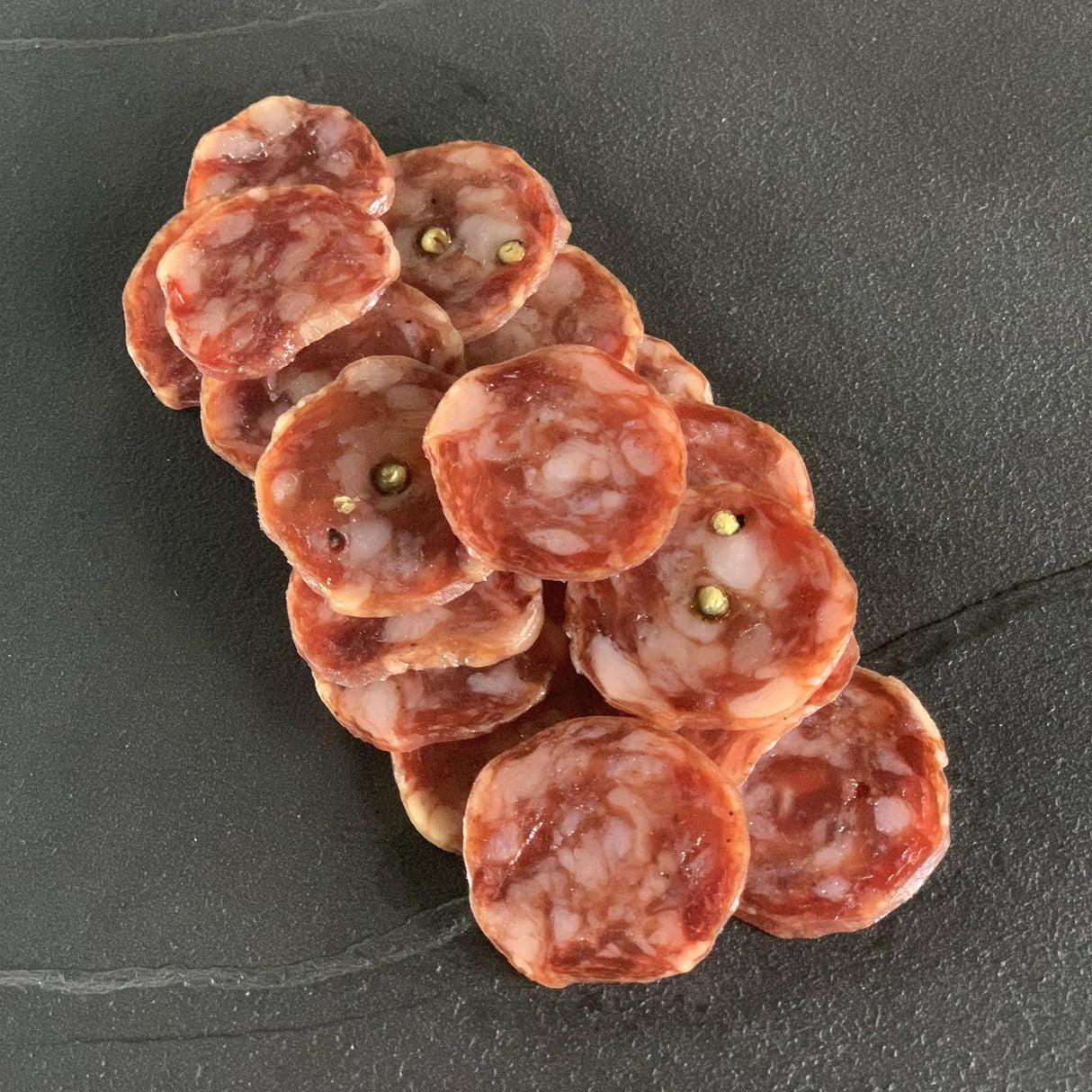 Sliced French salami arranged on a dark slate surface, showing visible peppercorns.