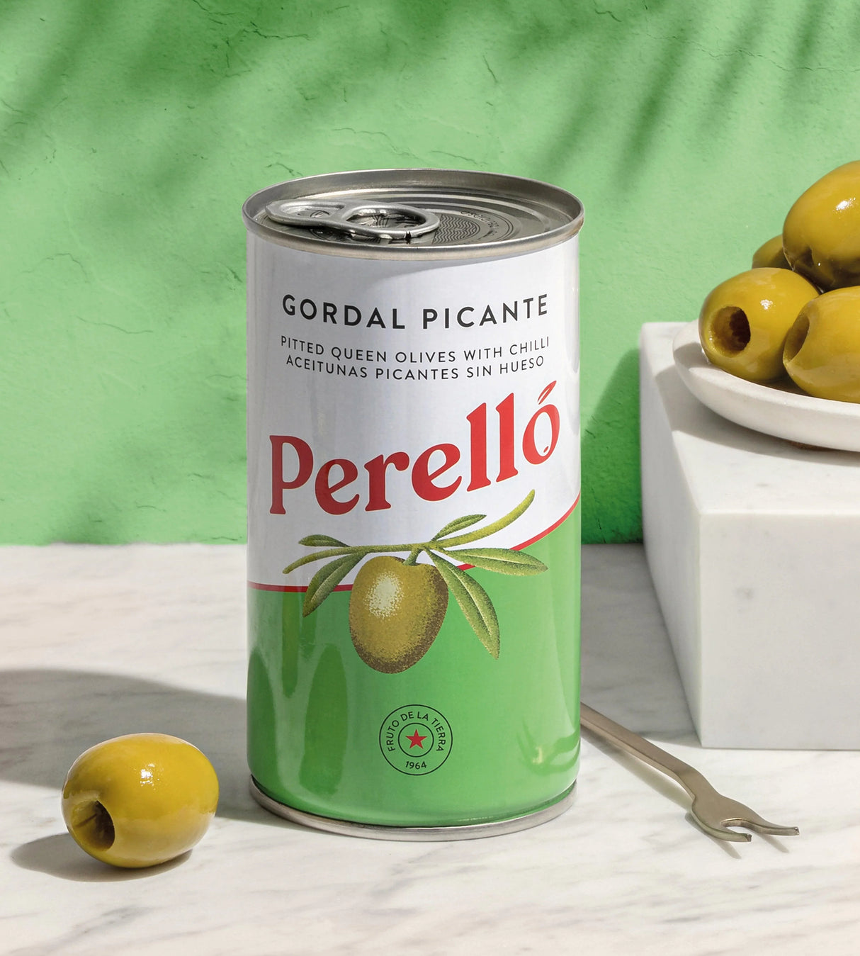Can of Perelló Gordal Picante pitted queen olives with chili on marble surface, whole olives beside.
