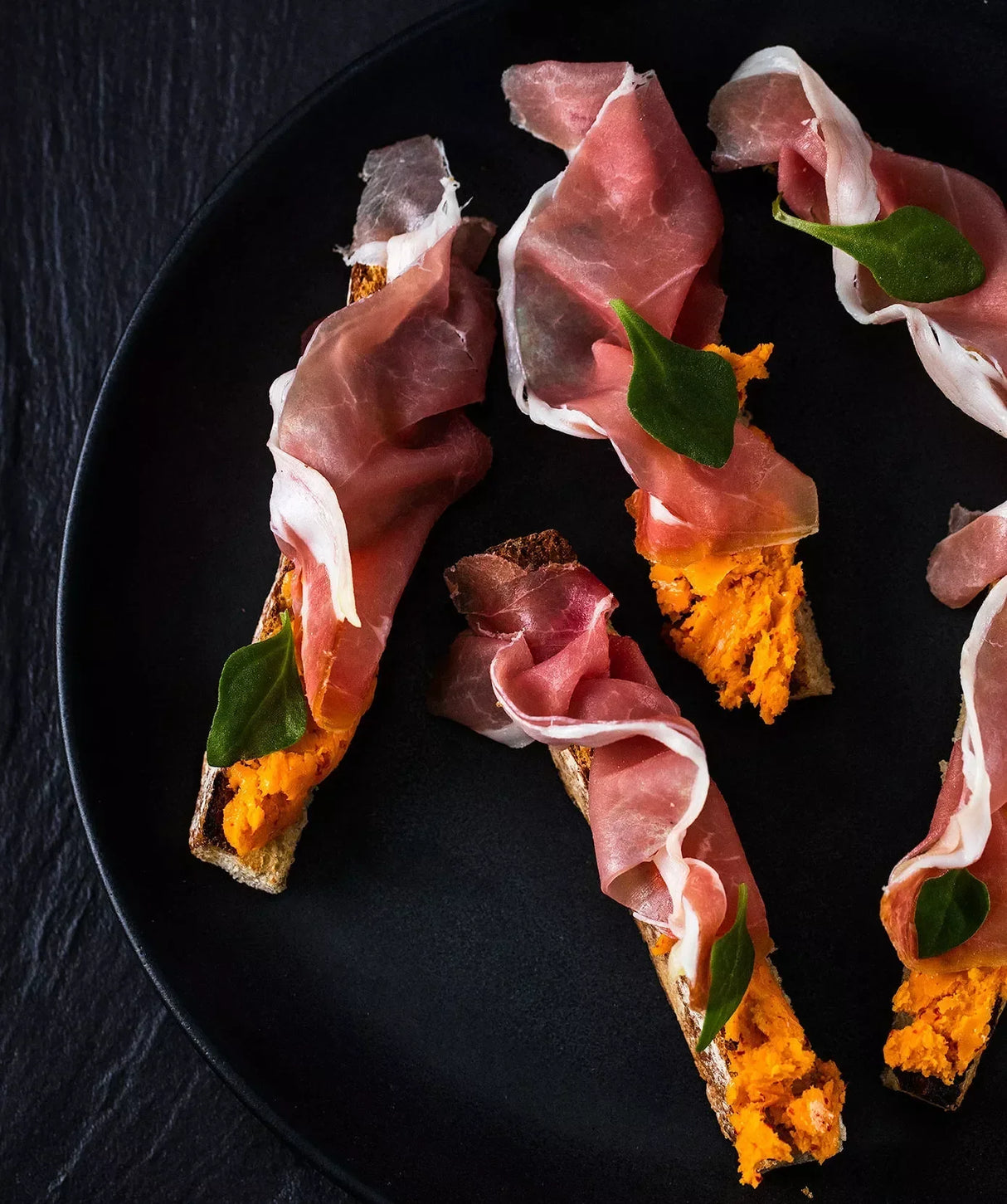 Artisan prosciutto crostini with orange spread and fresh basil on black plate