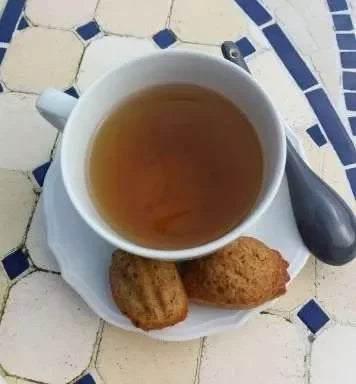 Cup of tea with two French madeleine cookies on a white plate, outdoor café table.
