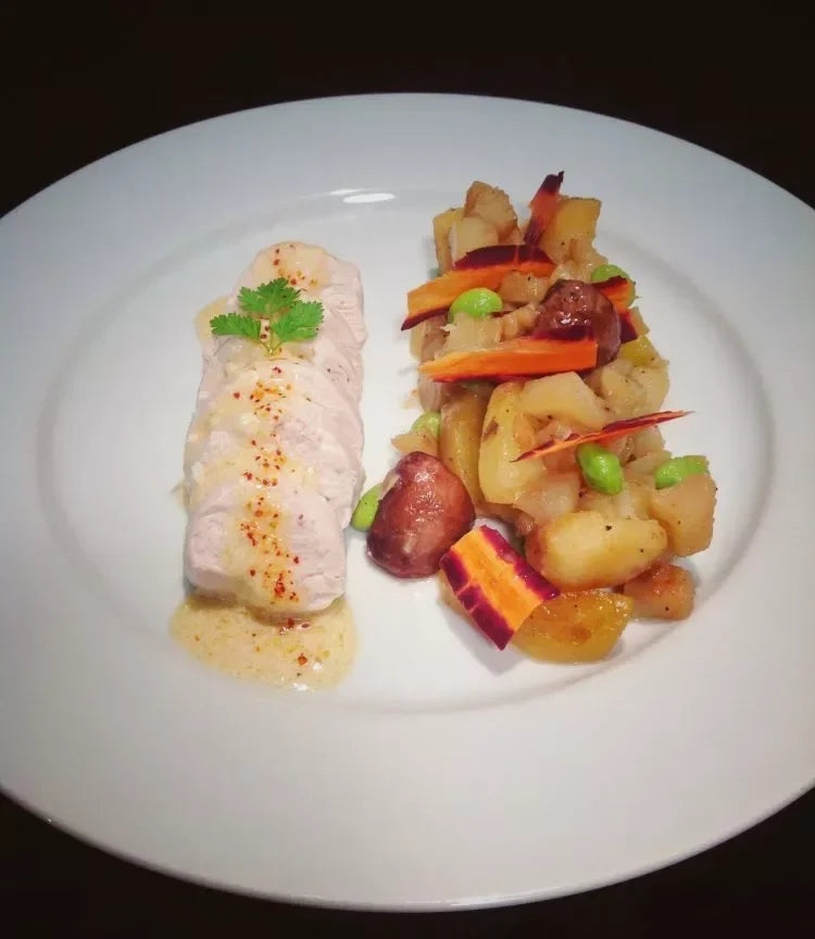 Sliced chicken breast with herb garnish, served with roasted potatoes, carrots, and edamame on a white plate.