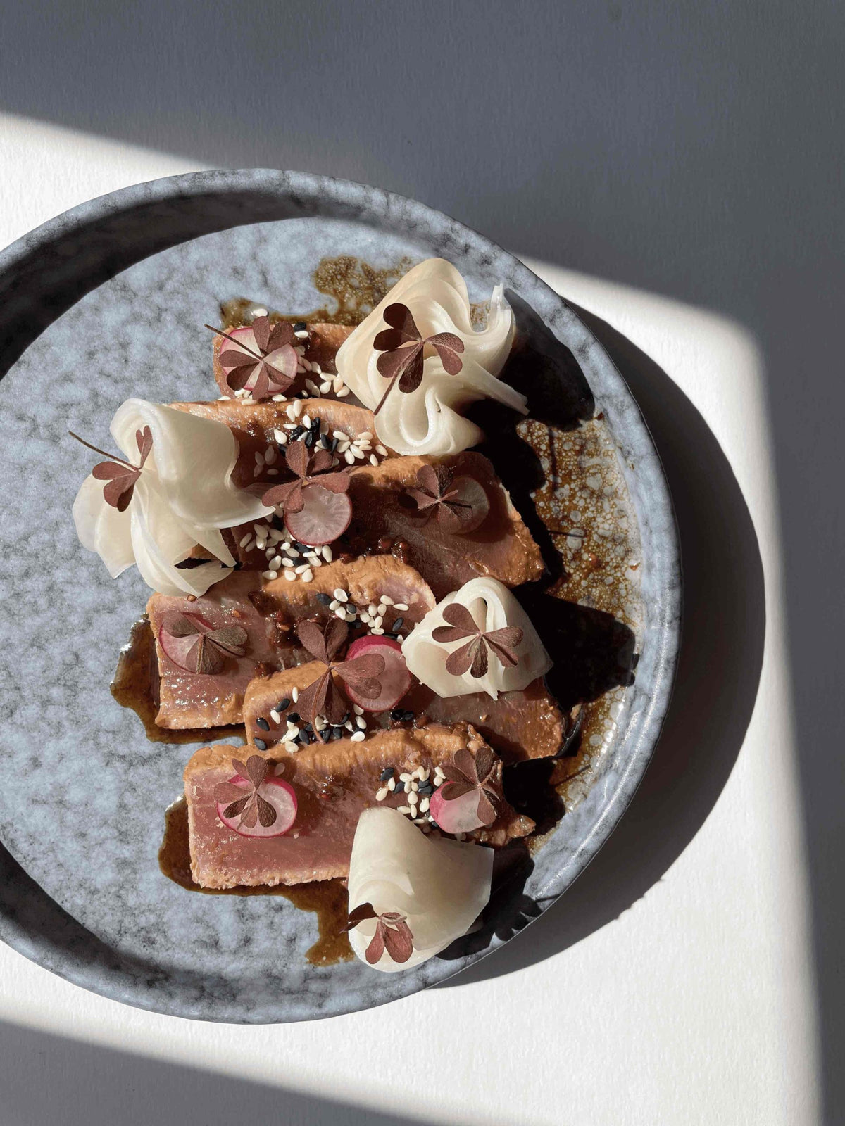 Slices of seared tuna tataki fanned on a plate with raspberry butter sauce, radish slices and sesame seeds