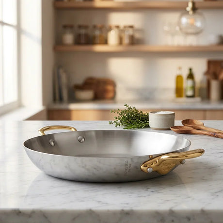 Mauviel stainless steel pan with gold handles on a white marble countertop.