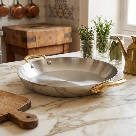 Large stainless steel paella pan with gold-tone handles on a marble countertop