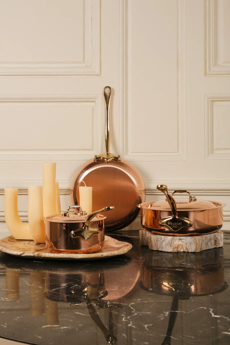 Mauviel copper cookware set with a frying pan, small saucepan, and lidded pot on a marble surface.