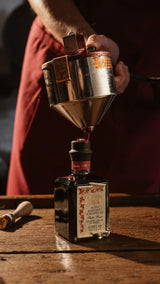 Person pouring liquid from a metallic container into a decorative bottle on a wooden surface.
