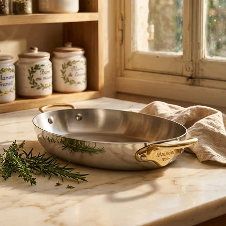 Stainless steel Mauviel roasting pan with gold handles on a marble countertop.