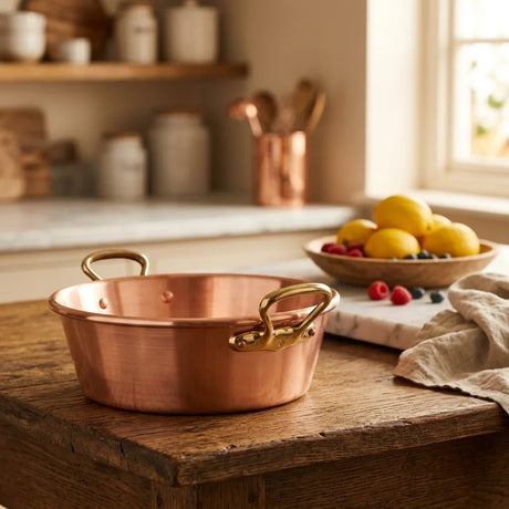 Copper saucepan with brass handles on a rustic wooden table near a bowl of lemons and berries.