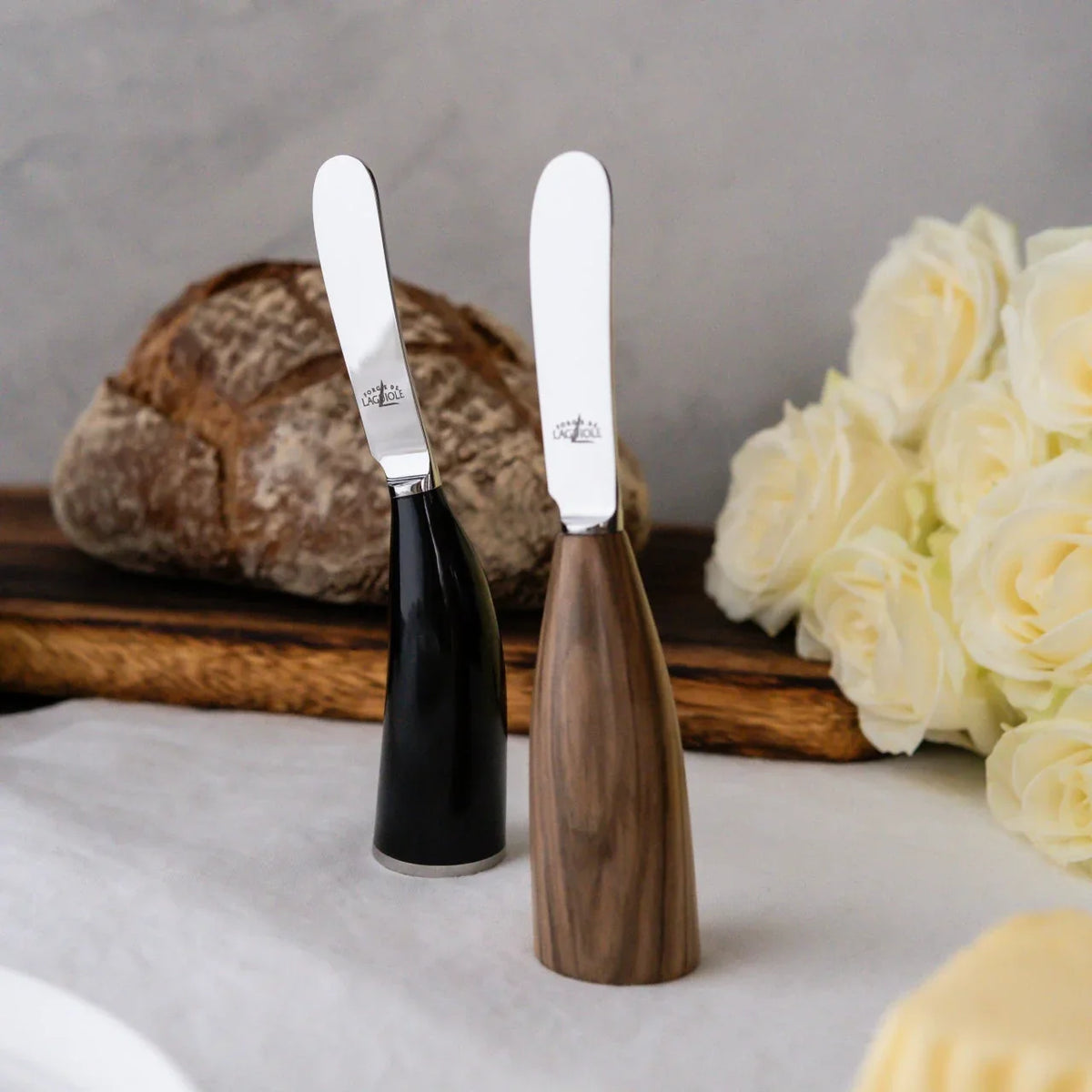 Two Laguiole butter knives with black and wooden handles standing on a white cloth.