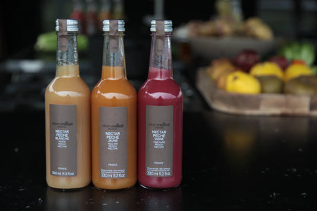 Three bottles of Alain Milliat French peach nectar on a black counter, fruit platter in background.