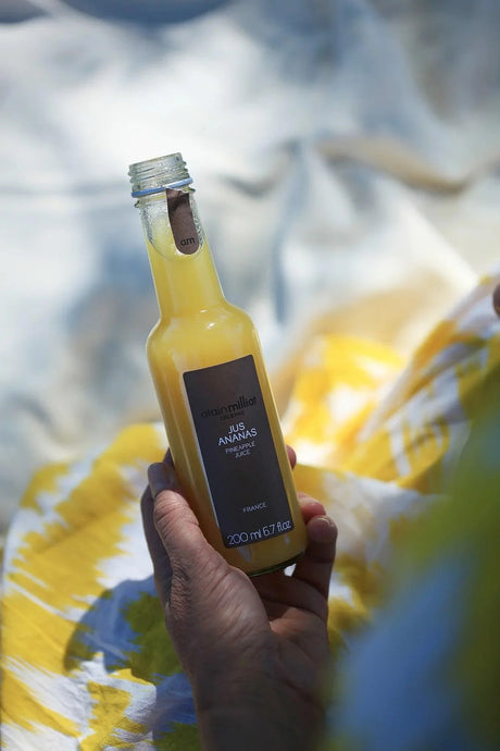 Hand holding Alain Milliat French pineapple juice bottle outdoors on yellow patterned fabric.