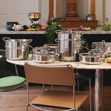 A collection of stainless steel pots and pans on a marble table with vegetables in the background.