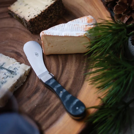 Assorted cheese wedges and a cheese spreader on a wooden board with pine branches.