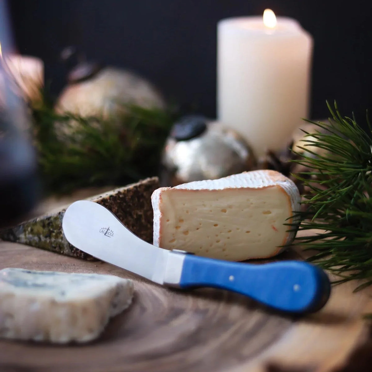 A wedge of soft white cheese and a blue-handled Laguiole cheese knife on a wooden board.