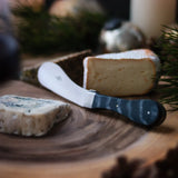 A wedge of blue cheese and a wheel of Brie cheese with a cheese knife on a wooden board.
