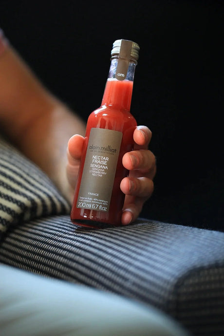 Hand holding Alain Milliat Sengana strawberry nectar bottle, premium French drink on striped fabric